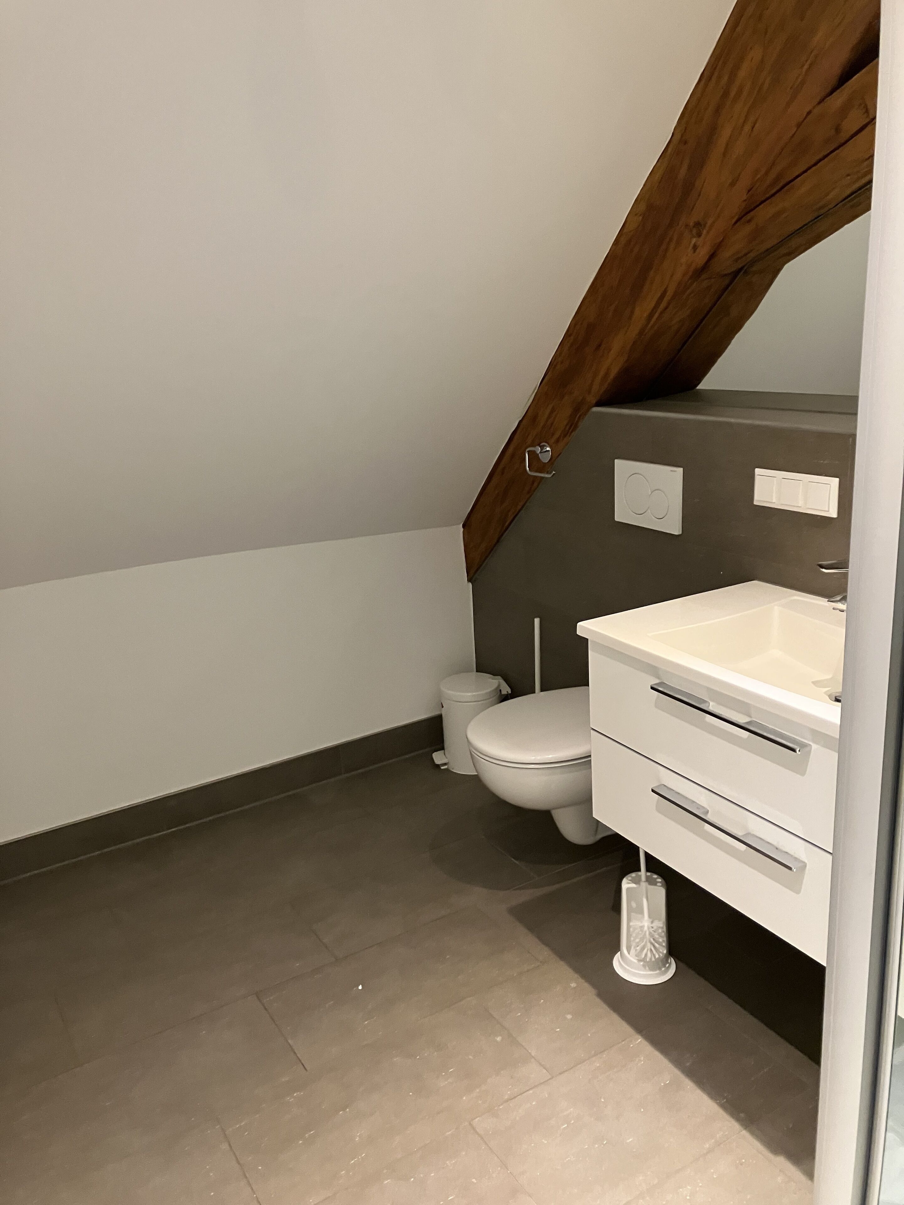 A small, modern bathroom with a white toilet, a white sink, and a mirror. There is a wooden beam above the sink, and the walls are painted white.