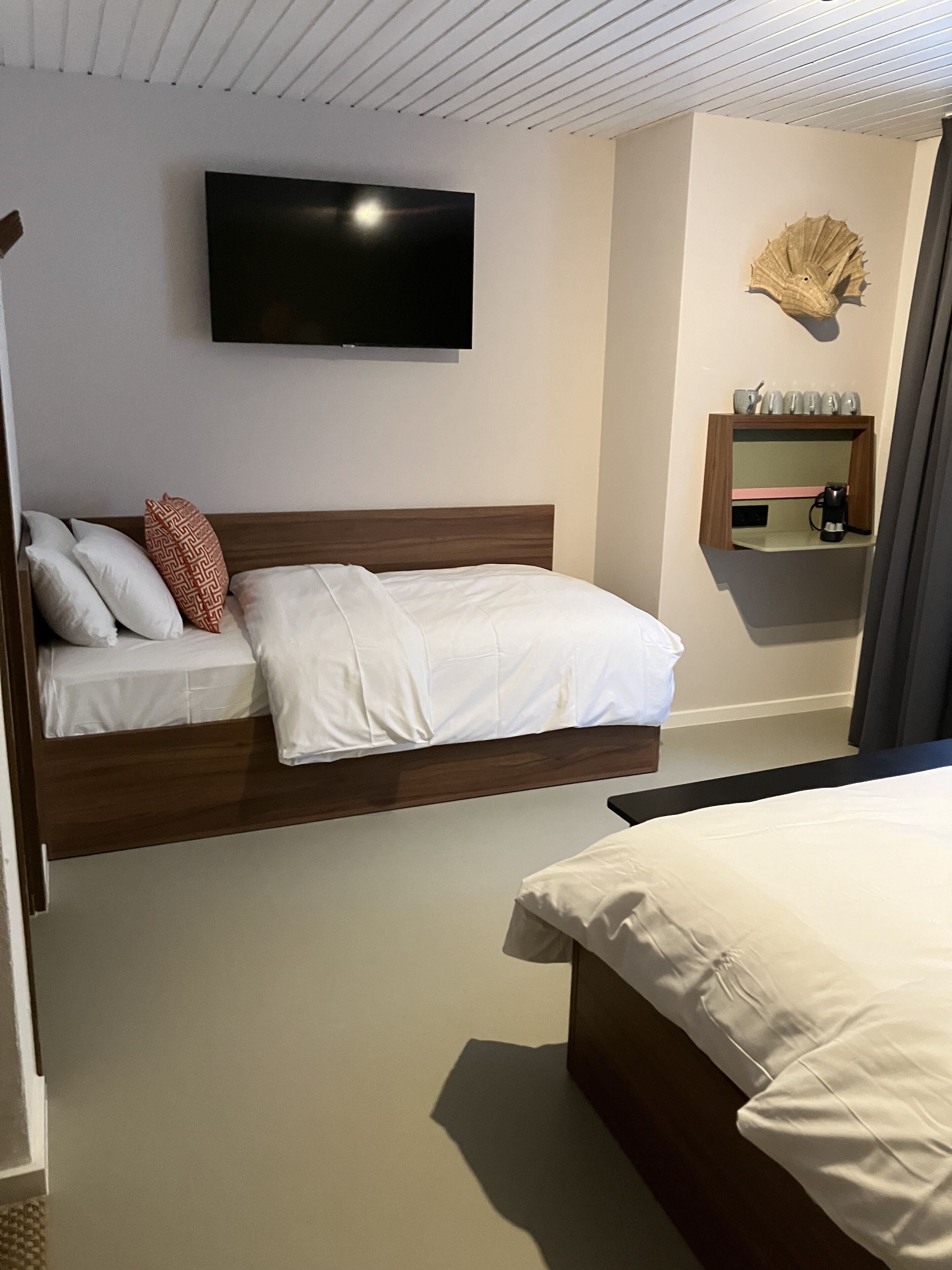 Een hotelkamer met een bed, een aan de muur bevestigde televisie, een klein plankje met een koffiezetapparaat en een decoratieve schelp aan de muur.