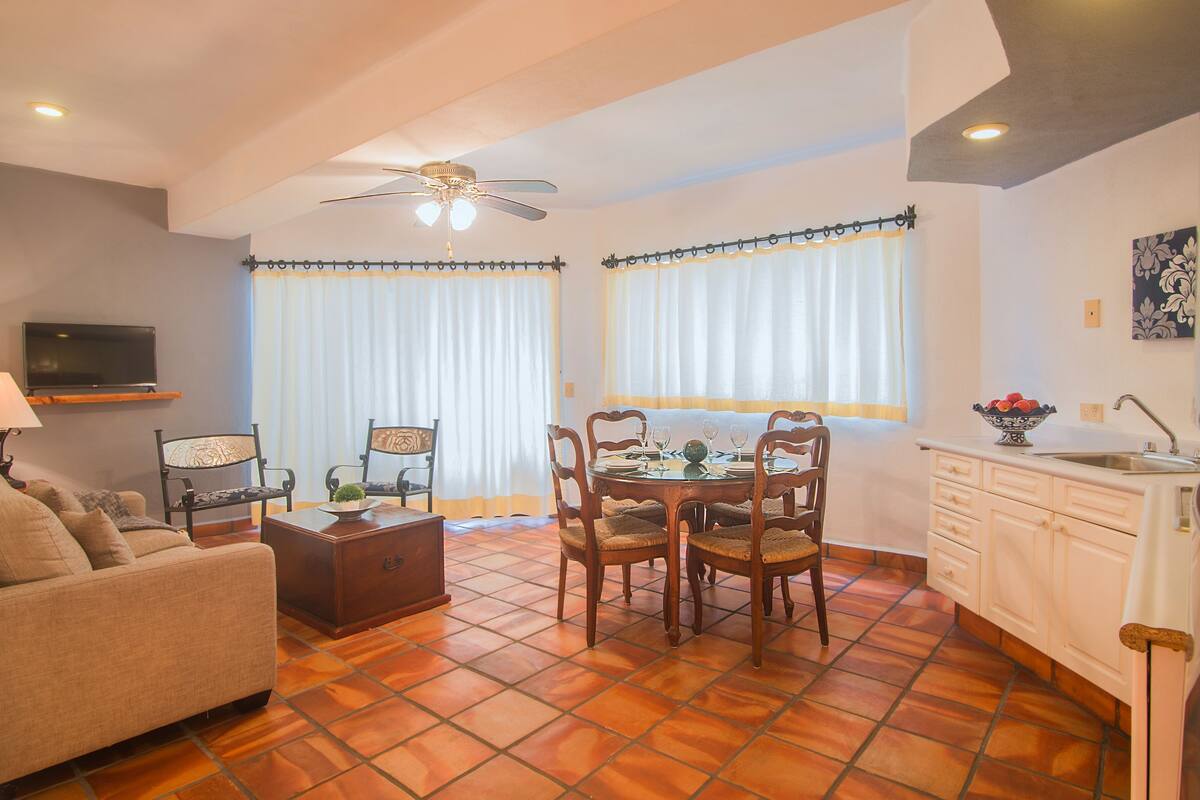 A living room with a sofa, a TV on a shelf, a dining table with chairs, and a kitchen area with a sink and cabinets.