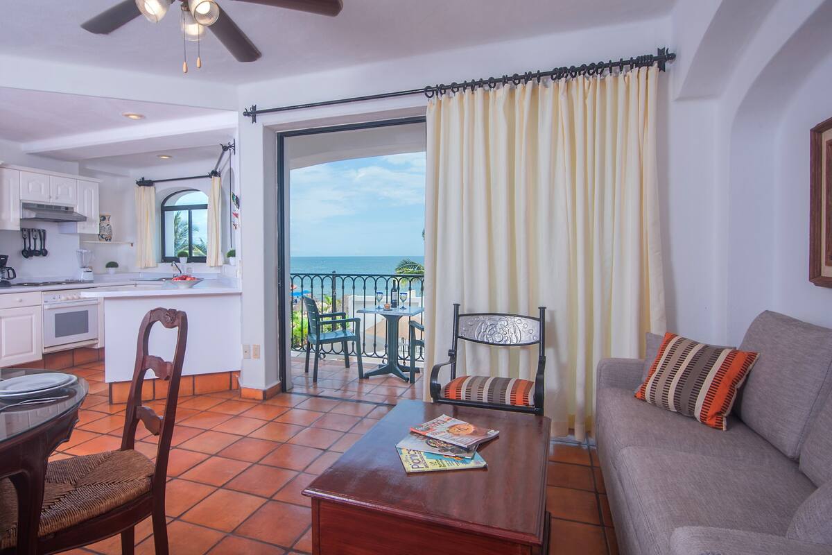 A modern kitchen with white cabinets, a dining area with a wooden table and chairs, a balcony with a view of the ocean, and a living room with a gray sofa and decorative pillows.