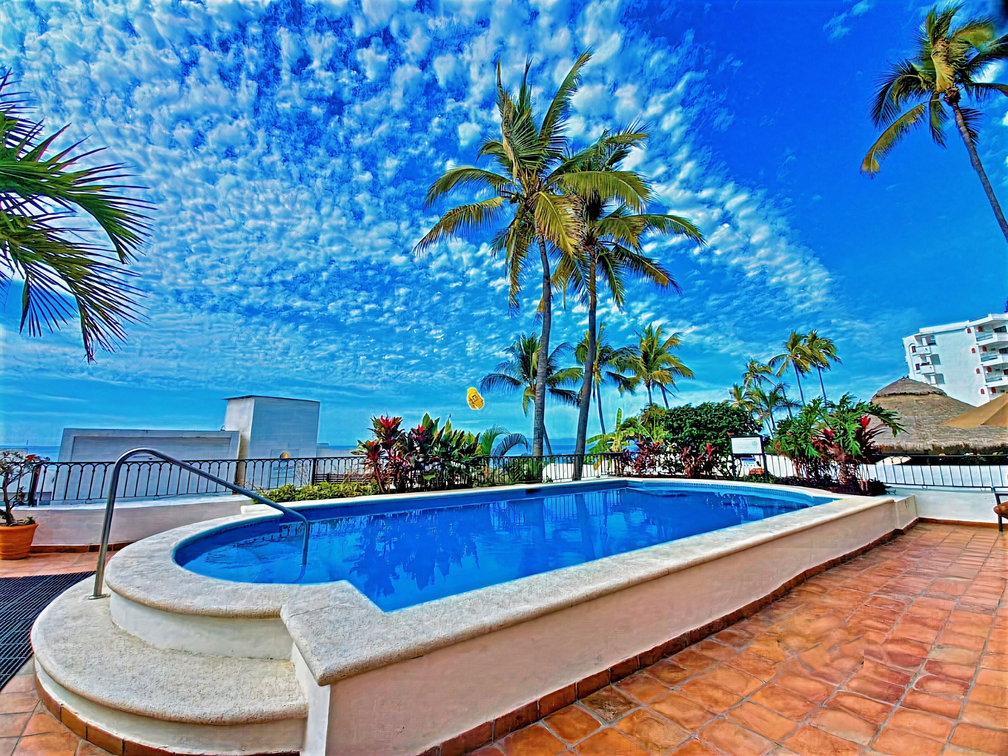 Ein Swimmingpool mit Steinrand, umgeben von Palmen und wolkenlosem, blauem Himmel.