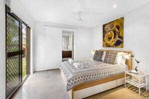 Relax in this serene bedroom with a large bed, ceiling fan, and floor lamp, offering a peaceful view of the surrounding greenery.
