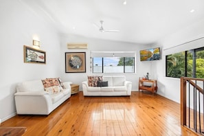Relax in this spacious living room with high ceilings and hardwood flooring, featuring two sofas, a side table, and large windows offering a view of the outdoors.