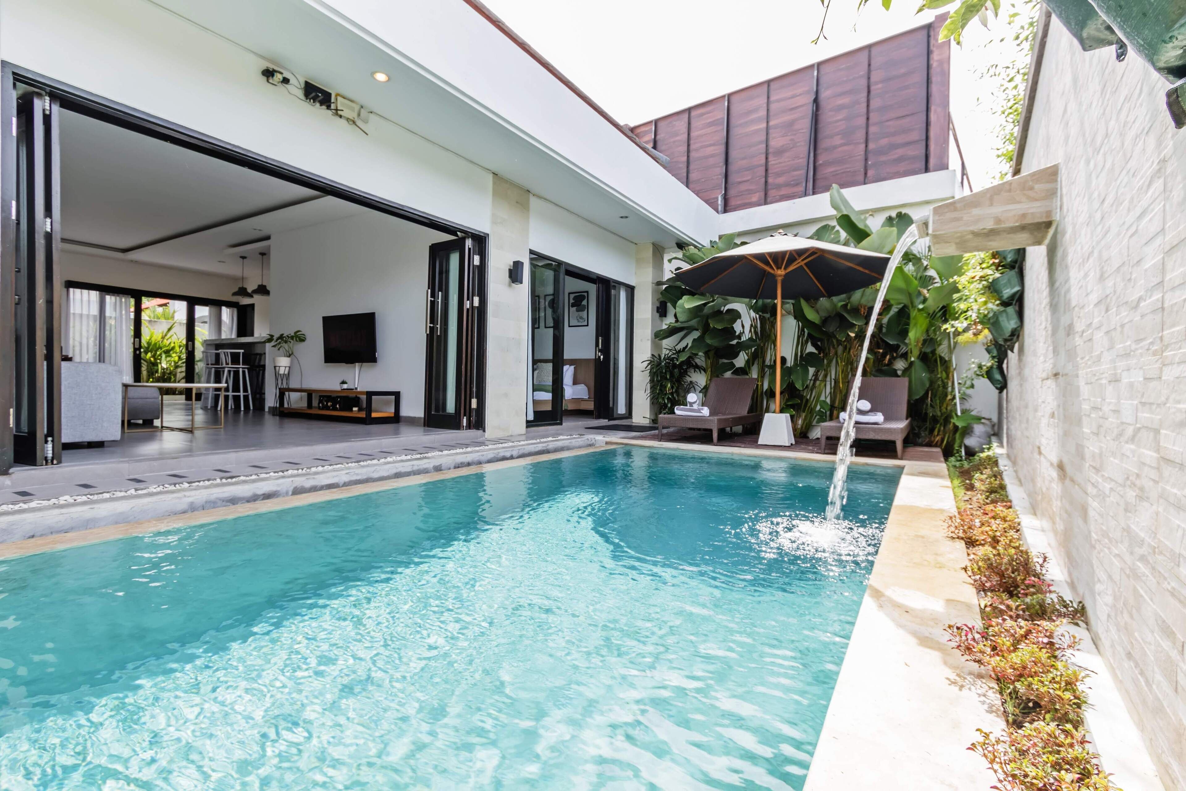 Una piscina moderna al aire libre con agua azul cristalina, rodeada por un patio con mobiliario y un gran paraguas.