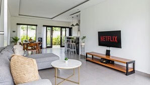 Ett modernt vardagsrum med en grå soffa, ett runt soffbord, ett TV-bord i trä och en stor TV med platt skärm som visar "NETFLIX".