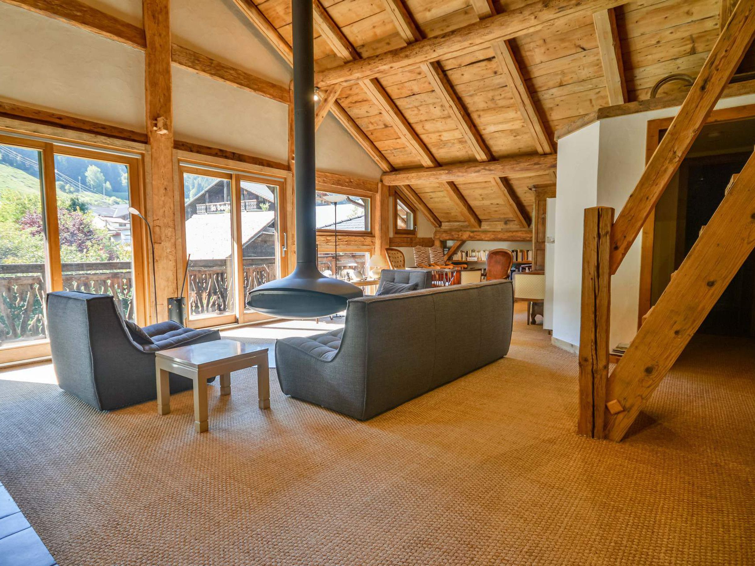 Amplia sala de estar con techo alto, vigas de madera, un sofá, una mesa y ventanas grandes que ofrecen vistas al exterior.