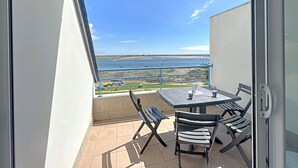 Un balcon avec une table et des chaises, donnant sur une plage où l’on aperçoit des bateaux et un ciel dégagé.