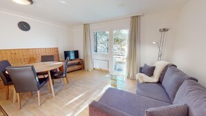 Ein modernes Wohnzimmer mit einem Holzesstisch, zwei Stühlen, einer Couch, einem Fernseher und einem großen Fenster mit Vorhängen.