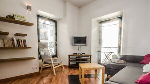 Una sala de estar moderna con un sofá gris, una mesa de centro de madera, un sillón gris y un televisor sobre un mueble. Se observan estanterías a la izquierda y una ventana con persianas a la derecha.
