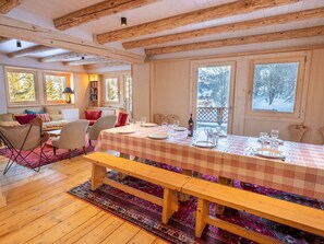 A cozy cabin interior with a dining table set for four, wooden beams on the ceiling, large windows with snowy views, and a comfortable seating area with a sofa and chairs. - Spacious chalet in Méribel with sunny terrace and fireplace (Méribel)