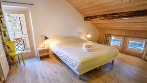 A bedroom with a wooden ceiling, a bed with white linens, a nightstand with a lamp, a chair, and a window with a view. - Spacious chalet in Méribel with sunny terrace and fireplace (Méribel)