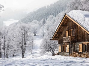 A wooden cabin with a balcony, surrounded by snow-covered trees and a mountainous backdrop.