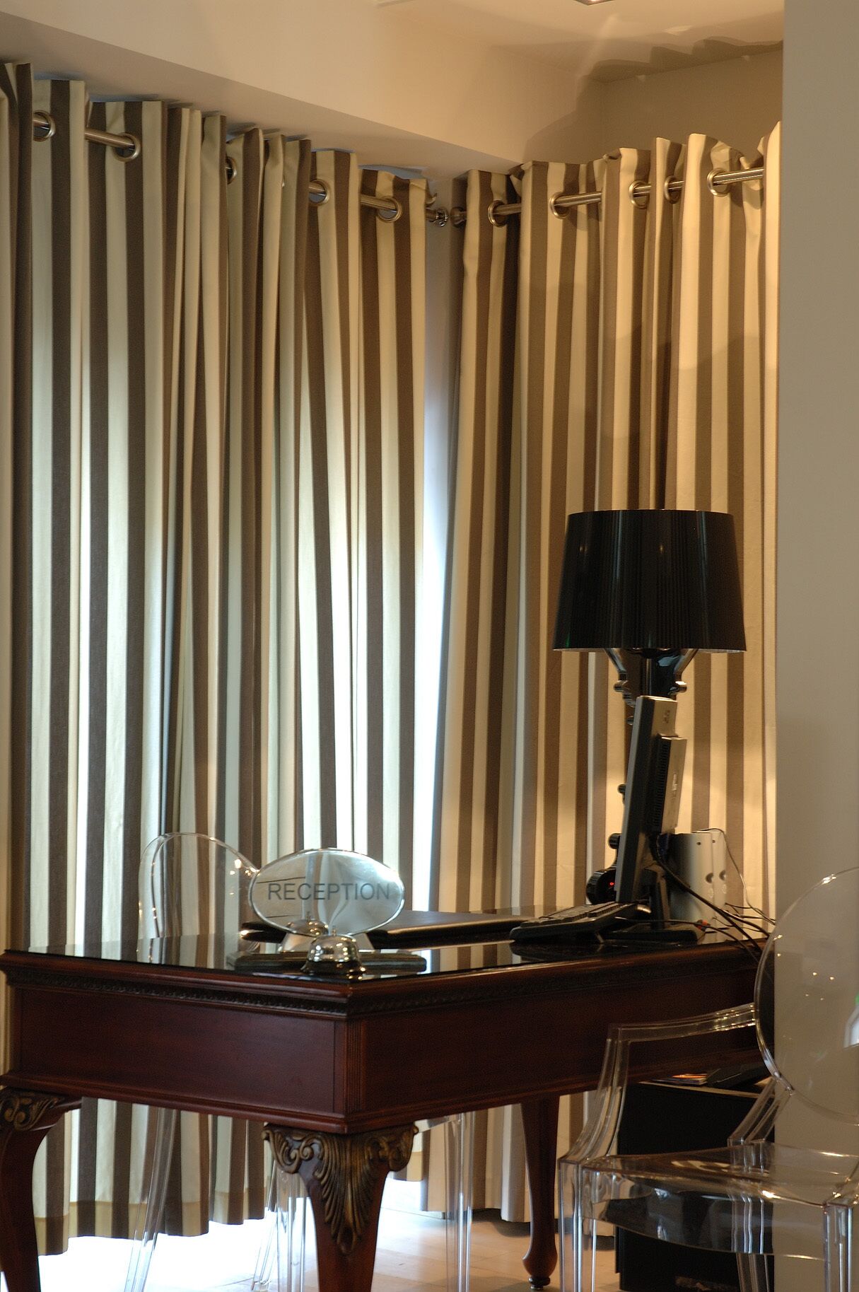 Une pièce avec un bureau, une lampe et une chaise. Il y a un rideau à rayures verticales.