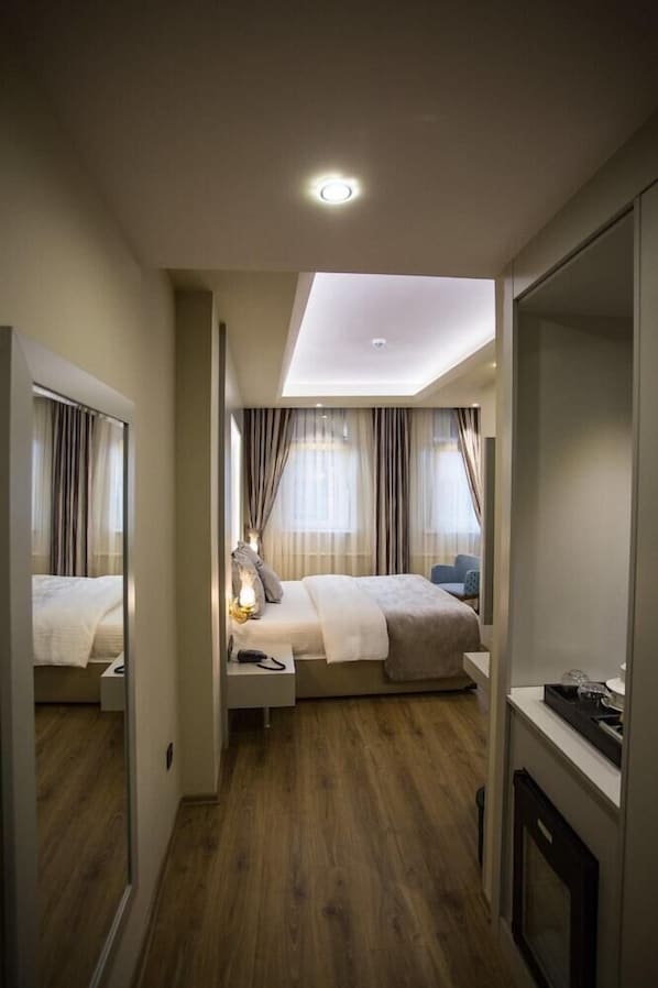 A hotel room with a wooden floor, a bed with a white headboard, a bedside table with a lamp, and a mirror.
