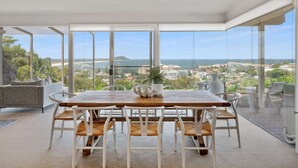 Ein moderner Essbereich mit Holztisch und weißen Stühlen, große Fenster mit Blick auf das Meer und die Stadt.
