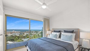 Ein modernes Schlafzimmer mit einem großen Bett, zwei Nachttischlampen, einem Deckenventilator und Blick auf eine Küstenstadt durch eine Schiebetür aus Glas.