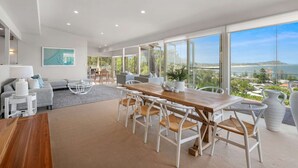 Ein modernes Wohnzimmer mit einem Holzesstisch, weißen Stühlen und einer Couch. Ein großes Fenster bietet Blick auf den Ozean und die Hügel.