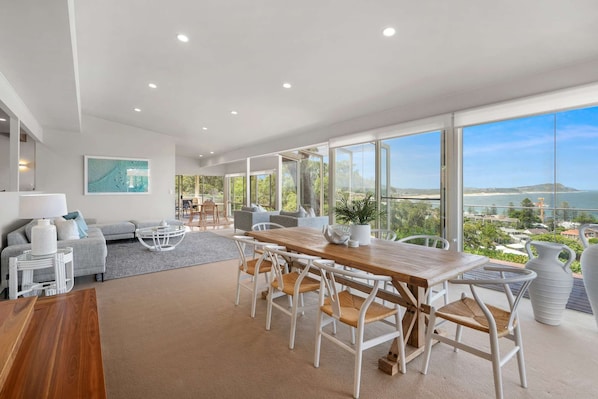 A modern living room with a wooden dining table, white chairs, and a sofa. There is a large window with a view of the ocean and hills.