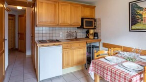A compact kitchen with wooden cabinets, a microwave, and a small refrigerator. A dining table with a checkered tablecloth is set for two.