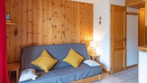 A room with a wooden wall, a bed with blue bedding and two yellow pillows, a bedside table with a lamp, and a wooden door.