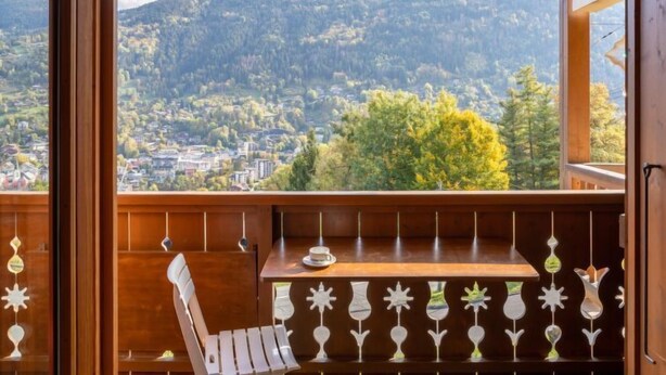 A balcony with a wooden chair and table, overlooking a mountainous landscape with a town below.