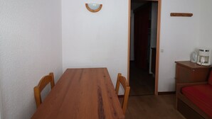Ein kleiner, schlichter Raum mit einem Holztisch und -stühlen, einem Bett mit roter Matratze und einer Kaffeemaschine auf einem Nachttisch.