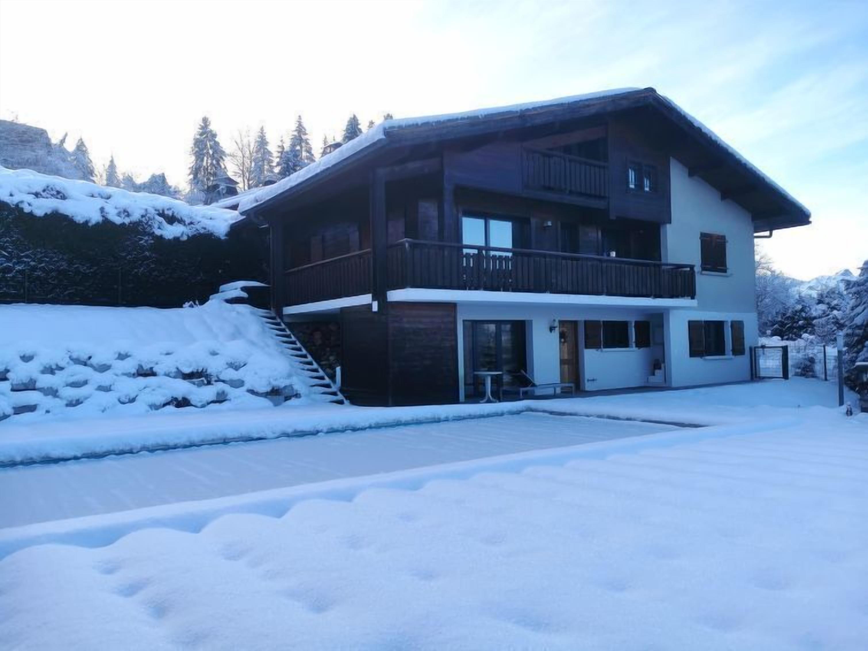 Un chalet de dos pisos con un balcón, un camino de entrada cubierto y un paisaje nevado.