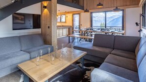 Un interior moderno con techo de madera, sofás grises, un comedor con mesas y sillas, y vistas a montañas a través de amplios ventanales.