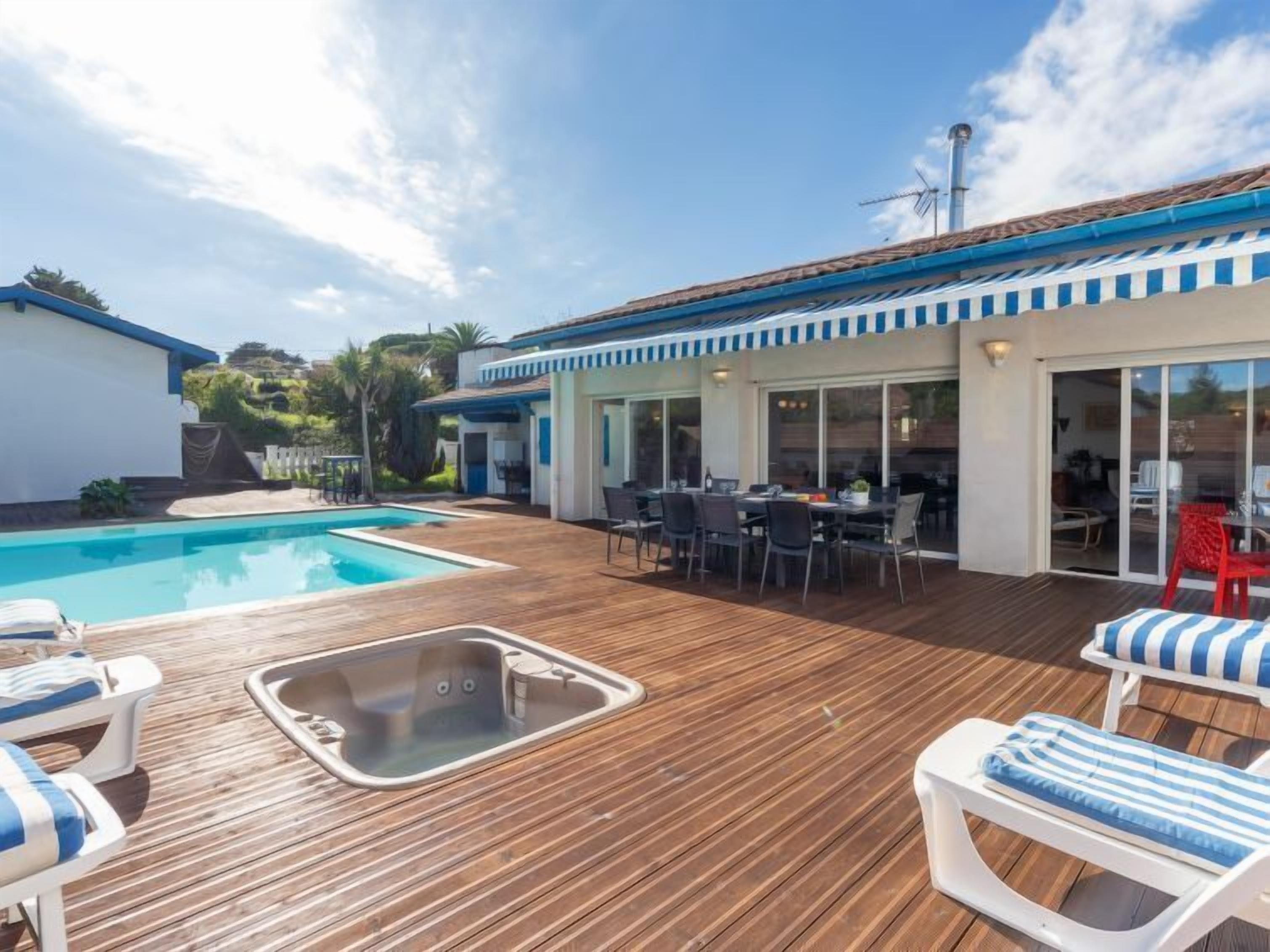 Un espace piscine doté d’une terrasse en bois, de chaises longues, d’un coin repas et d’un patio couvert avec une table et des chaises.
