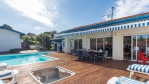 Een zwembadgedeelte met een houten terras, ligbedden, een eethoek, en een overdekte patio met een tafel en stoelen.