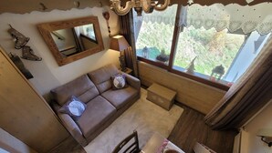 A cozy living area with a brown sofa, a small table with a candle, and a window with a view of greenery.