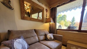 A brown leather sofa with patterned cushions, a wooden-framed mirror, a lamp, and a window with a view of trees and a gazebo.