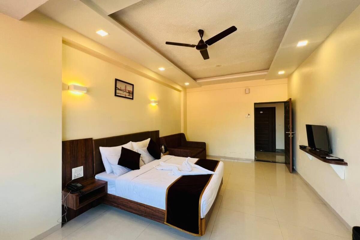 A hotel room with a bed, bedside table, wall-mounted TV, ceiling fan, and recessed lighting.