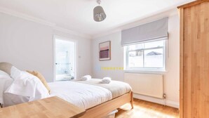 A bedroom with a bed, wooden bedside table, window with blinds, and a radiator. - 2 Bed Apt| Parking| Central Windsor By Tempstay (Windsor)