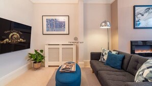 A modern living room with a sofa, ottoman, fireplace, framed artwork, and a radiator. - Charming Character Home| 1 Bed| By Tempstay (Windsor)