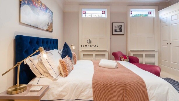 A bedroom with a large bed, a desk with a lamp, a painting on the wall, and a sign that says 'TEMPSTAY'. - Charming Character Home| 1 Bed| By Tempstay (Windsor)