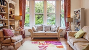 A cozy living room with a beige sofa, two armchairs, a coffee table, and a bookshelf. Large windows offer a view of greenery outside. - Luxury Home- Parking- Walk 2 Sights By Tempstay (Windsor)