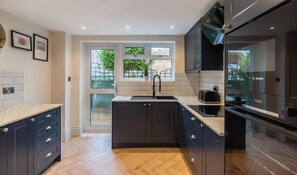 A modern kitchen with dark cabinetry, a built-in sink, and a window with a view of greenery. - Modern Windsor 4 Bed Home W/ Parking - By Tempstay (Windsor)