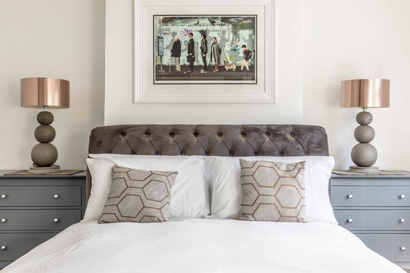 A bedroom with a tufted headboard, two bedside tables with lamps, a framed artwork above the bed, and a bed with patterned pillows.
