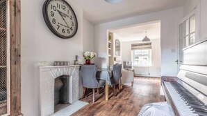 A room with a piano, a fireplace, a dining table with chairs, and a large wall clock. - Stunning Victorian Cottage in Windsor By Tempstay (Windsor)