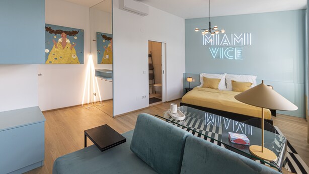 Une chambre d’hôtel moderne, équipée d’un lit, d’un canapé, d’un bureau et d’un panneau néon sur lequel on peut lire « MIAMI VICE ».