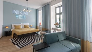 Une chambre d’hôtel moderne, équipée d’un lit, d’un canapé, d’un bureau et d’un panneau lumineux néon sur lequel on peut lire « MIAMI VICE ».