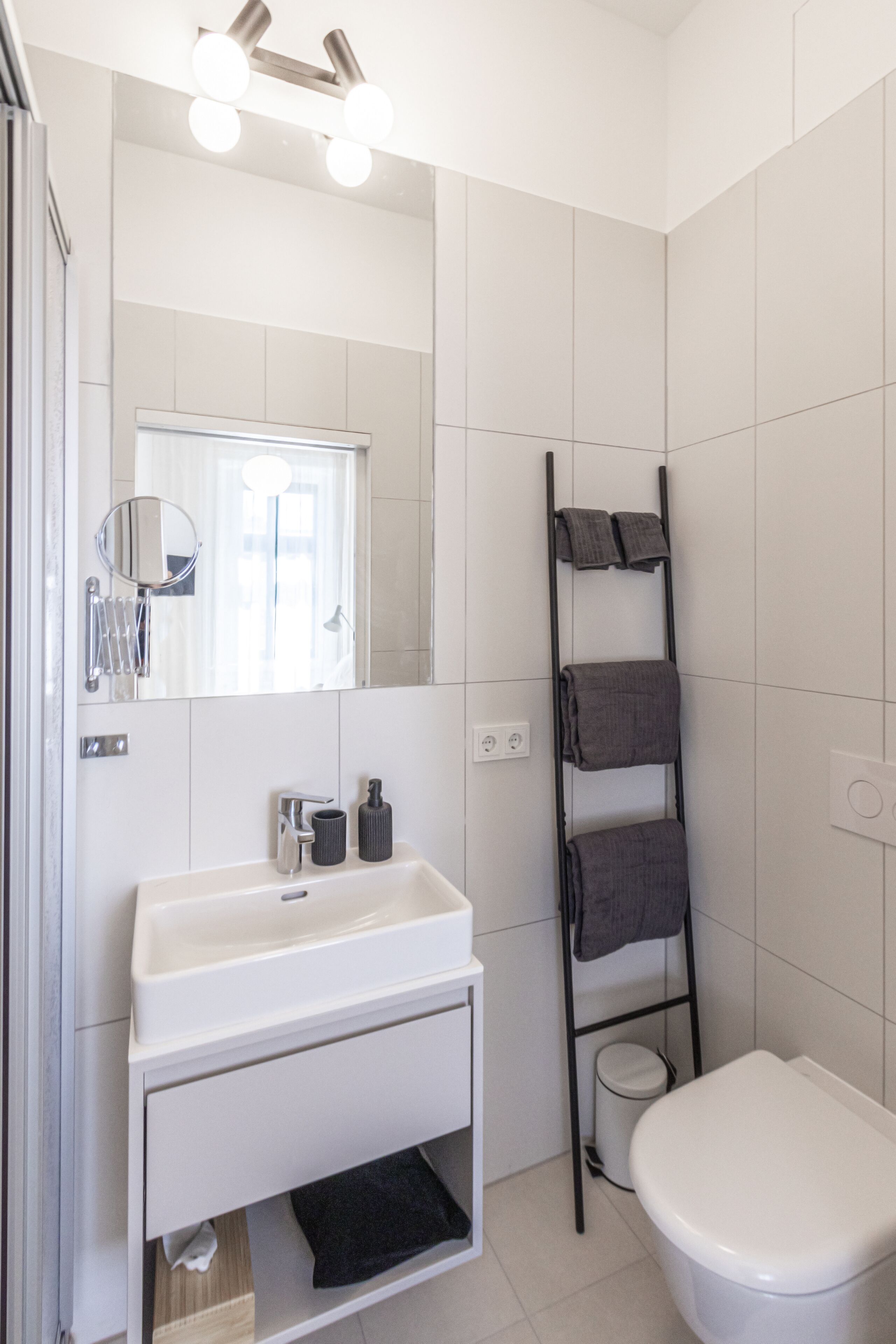 A bathroom with a white sink, mirror, and toilet. There is a towel rack with towels and a trash bin.