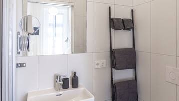 Baño con lavamanos blanco, espejo e inodoro. Se observa un perchero con toallas y un cesto de basura.