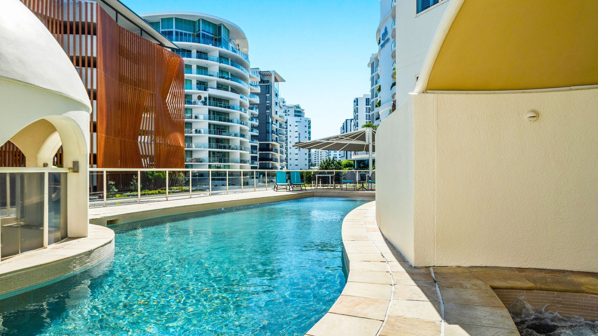 A swimming pool with a curved design, surrounded by modern buildings with balconies and a clear sky.