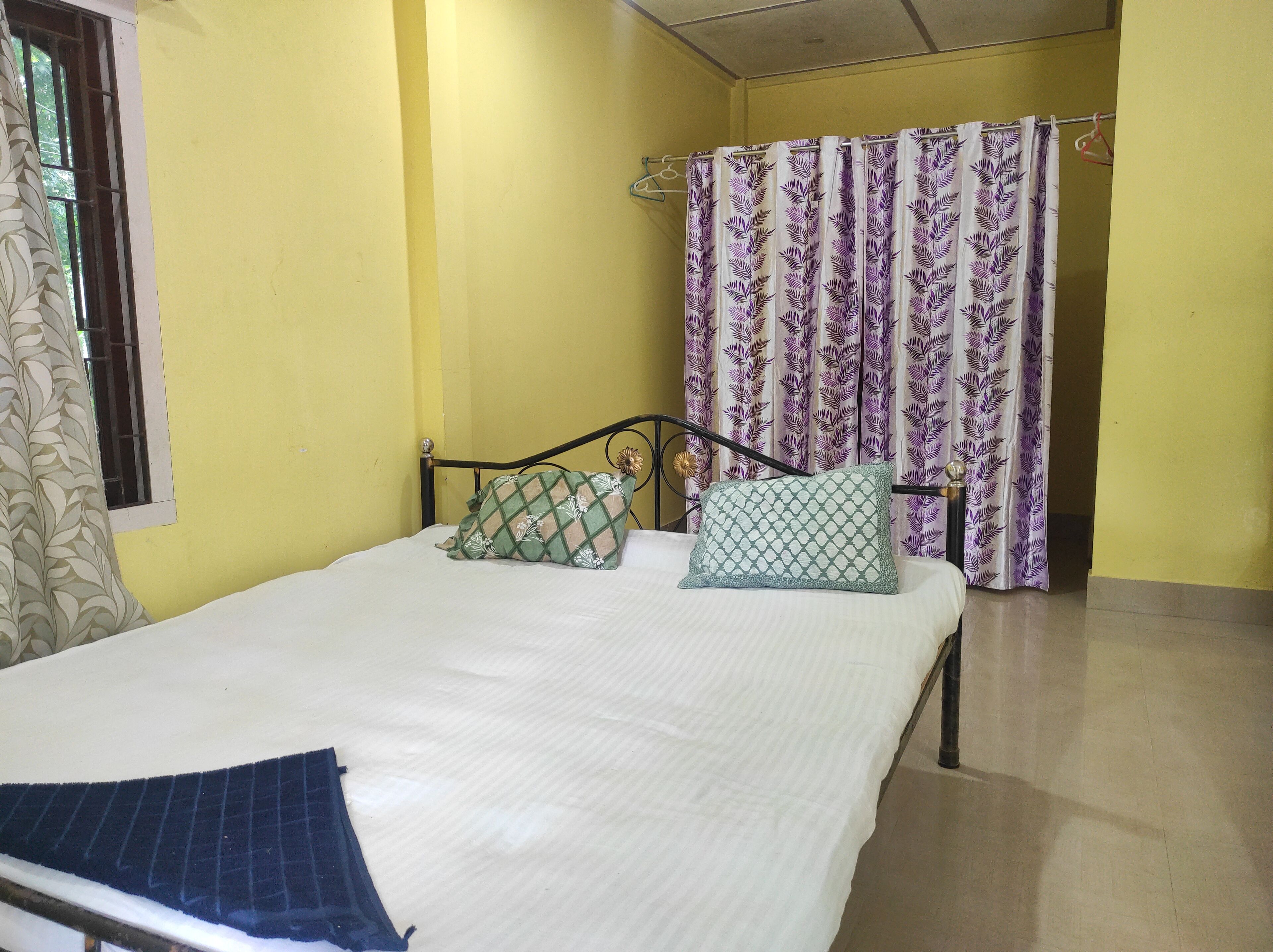 A single bed with two pillows, a window with a curtain, and a shower curtain with a leaf pattern.