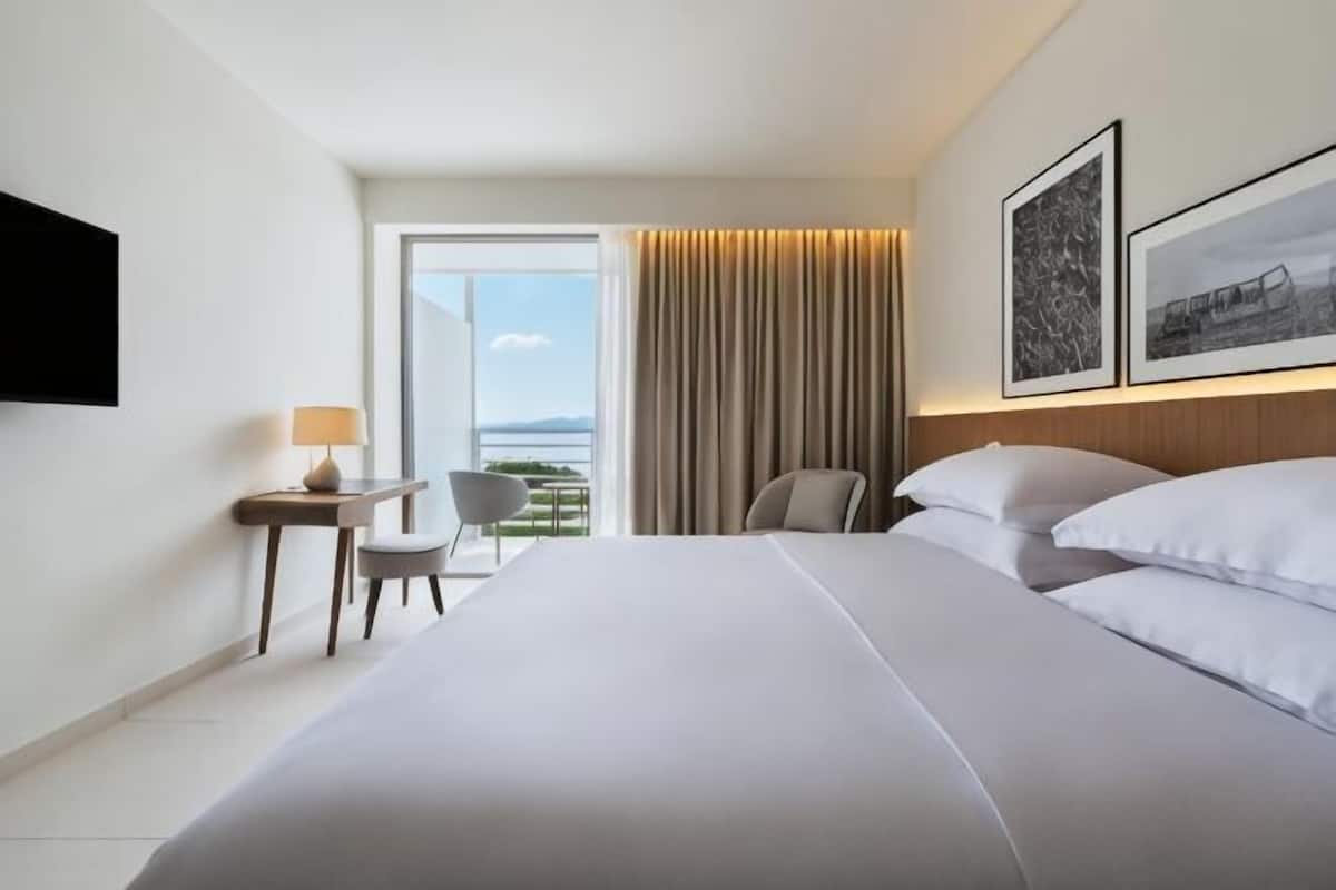 Une chambre d’hôtel avec un grand lit, un bureau avec une chaise, une télévision et un balcon avec des rideaux.