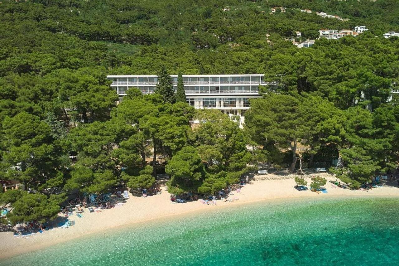 Μια αεροφωτογραφία ενός παραθαλάσσιου ακιν ήτου, με ένα σύγχρονο κτίριο που περιβάλλεται από πυκνή βλάστηση, μια αμμώδη παραλία και κρυστάλλινα, γαλαζοπράσινα νερά.