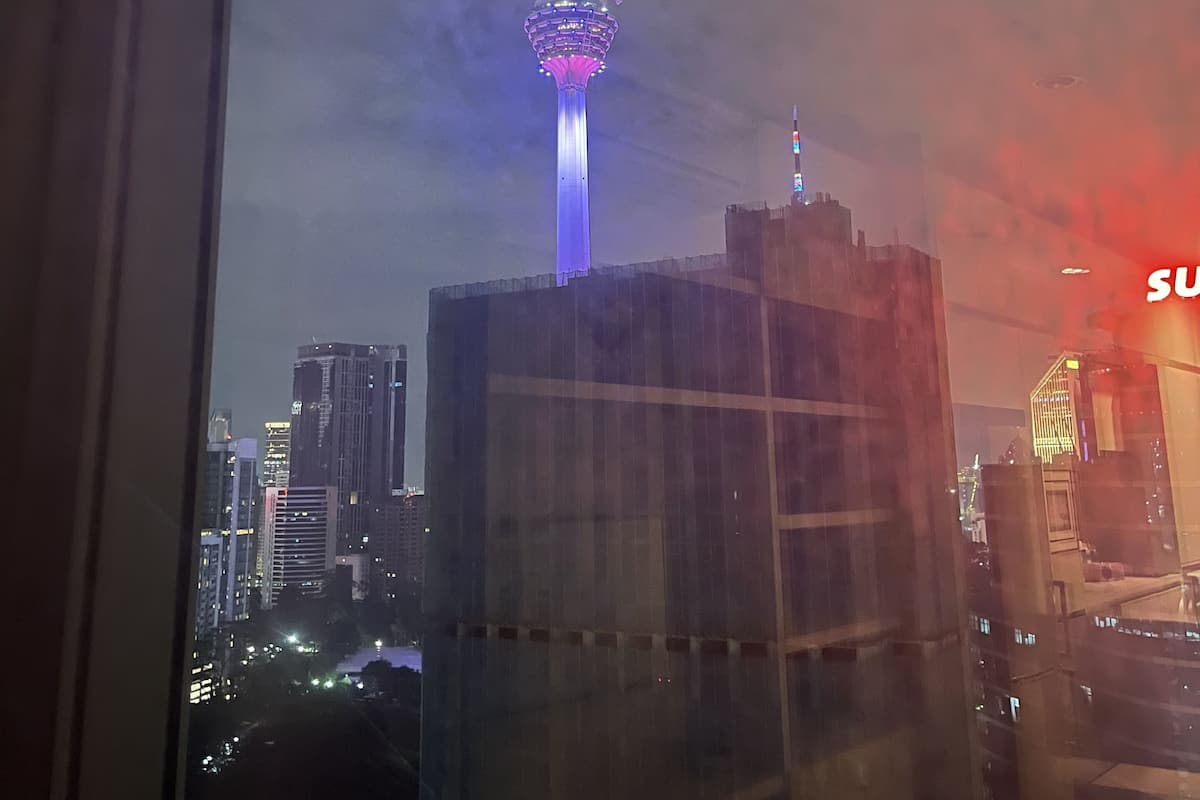 A cityscape with a prominent tower featuring illuminated rings and a spire, viewed through a window with a reflection of a building.
