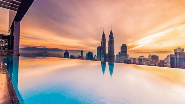 Blick auf eine Stadtlandschaft, wobei die Petronas Towers im ruhigen Wasser, vermutlich einem Pool, deutlich reflektiert werden. Ein modernes Gebäude ist links im Bild zu sehen.